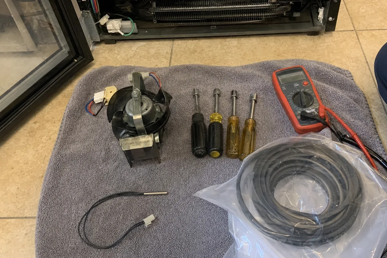 Meter, drivers and bagged replacement parts set out on a towel in front of an appliance opened for a sealed-system check